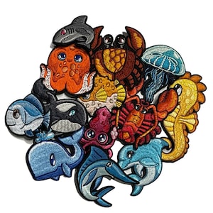 Può includere: Una collezione di toppe ricamate raffiguranti varie creature marine. Le toppe includono uno squalo, un polpo, un granchio, una medusa, un cavalluccio marino, un pesce, una balena, un'orca e un delfino. Ogni toppa è colorata con cuciture dettagliate.