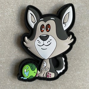 May include: A cartoon wolf patch with a black border. The wolf has grey, white, and black fur, red eyes, and a smiling expression. It sits beside a green ball with blue details. The patch is designed in a cartoon style.