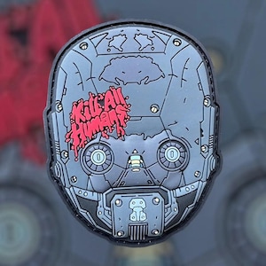 Op de afbeelding: Grijze patch in de vorm van een robothoofd met de rode tekst "Kill All Humans". Het ontwerp bevat mechanische details en een zwarte rand, geschikt voor kleding of tassen.