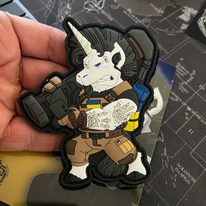 以下が含まれることがあります： 武器を持った漫画のユニコーン兵士を描いた、黒、白、タンのパッチ。ユニコーンは白い角、黒いたてがみを持っており、ウクライナの国旗のパッチが付いた軍服を着ています。パッチには黒い縁取りがあります。