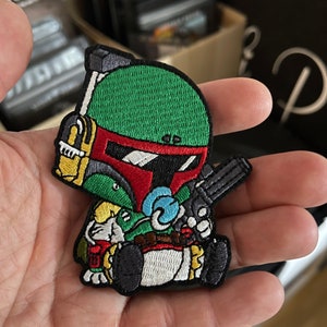 May include: Embroidered patch featuring a cartoon version of a character in a green helmet with red and white accents, holding a gun. The patch has a green, red, yellow, and blue color scheme. The patch is in the shape of the character.