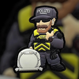 Könnte beinhalten: Ein Cartoon-Patch mit einem Polizisten in grauer Uniform, Sonnenbrille und Kappe mit dem Text "POLIZEI Assistenz". Der Beamte hält einen weißen Gegenstand mit silbernem Rand. Gelbe Akzente sind auf der Uniform zu sehen.