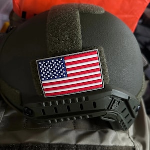 May include: A green tactical helmet with an American flag patch attached to the side. The helmet has a black strap and a black and green adjustable chin strap.
