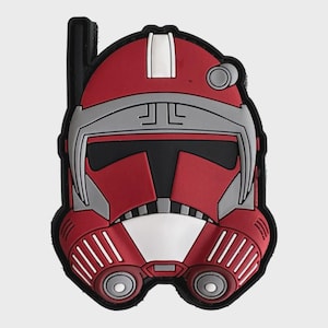 Könnte beinhalten: Ein rot, weiß, schwarz und grau geformter Aufnäher in Form eines Helms. Der Aufnäher zeigt ein detailliertes Design eines Helms mit Visier und anderen Details. Der Aufnäher hat einen schwarzen Rand und eine kleine schwarze Lasche oben links.