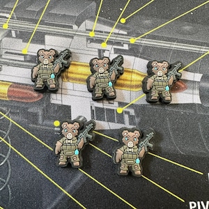 May include: Five black and brown bear-shaped shoe charms. Each charm depicts a bear wearing a military vest and holding a rifle. The charms are on a dark background with yellow lines and bullet casings.