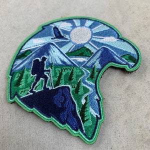 Op de afbeelding: Geborduurde patch in de vorm van een adelaarskop, met een wandelaar op een berg, een rivier, bomen en een zon. De patch is voornamelijk blauw en groen, met een groene rand. Het ontwerp roept een gevoel van avontuur en de buitenlucht op.