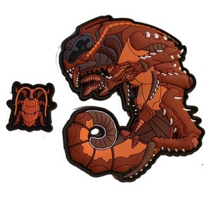 May include: Two rubber patches featuring insect-like creatures. The larger patch is a detailed, brown and orange design with sharp teeth and a curled tail. The smaller patch is a simplified, orange insect with a black border.
