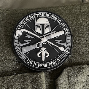Könnte beinhalten: Ein schwarzer und weißer runder Patch mit einem mandalorianischen Symbol, einem Boba Fett-Helm und einem Blastergewehr. Der Patch hat einen weißen Rand mit Text um den Rand.