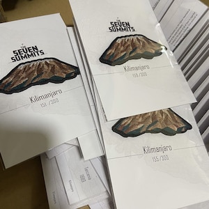 May include: Multiple cards featuring embroidered patches of Mount Kilimanjaro, part of the Seven Summits series. Each card displays the mountain's name and a unique number out of 300. The patches are brown and green.