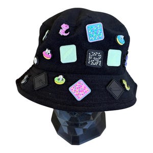 May include: Black bucket hat adorned with colorful, square-shaped patches and various small, decorative pins. The patches feature designs like sprinkles, a knife, and abstract patterns. The hat is displayed on a black geometric stand.