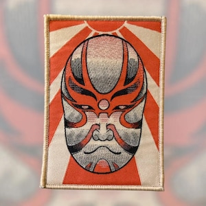May include: Embroidered patch featuring a stylized Kabuki mask in red, gray, and black, set against a background of red and white rays. The patch has a rectangular shape with a beige border.