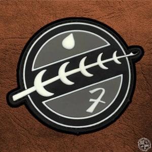 May include: A circular patch featuring a black and gray design with a white outline. The patch includes a stylized white spear, a drop shape, and the number 7. The background is a textured brown.
