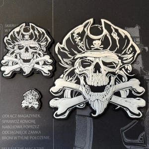 Peut inclure: Trois patchs brodés blancs représentant un crâne portant un chapeau de pirate et tenant des os croisés. Les patchs sont de tailles différentes et sont sur un fond noir.
