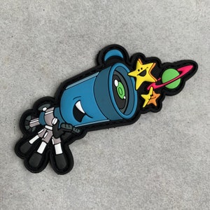 Może przedstawiać: Niebieska naszywka z teleskopem z kreskówki z uśmiechniętą twarzą, strzelająca żółtymi i pomarańczowymi gwiazdami oraz zieloną planetą z różowym pierścieniem. Naszywka ma czarną obwódkę i znajduje się na szarej powierzchni.