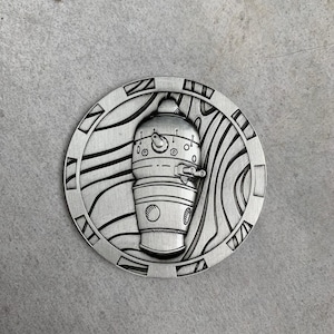 May include: A silver-colored, circular pendant with a detailed, engraved design. The central image is a stylized, cylindrical figure with various circular and rectangular details. The pendant is surrounded by a patterned border with geometric shapes and wavy lines.