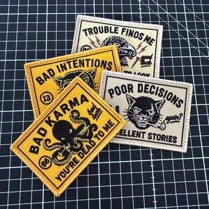 May include: A collection of four rectangular embroidered patches featuring black text and illustrations on yellow and off-white backgrounds. The patches display phrases including Trouble Finds Me, Bad Intentions, Bad Karma, and Poor Decisions with graphic icons.