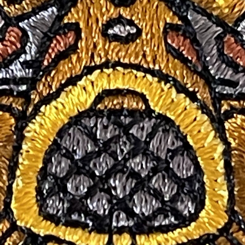 May include: Close-up of a black and gray embroidered design with a honeycomb pattern. The design is surrounded by a yellow and brown embroidered border.