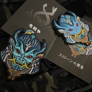 May include: Two embroidered patches with a blue and teal Oni mask design, red eyes, and detailed features. The patches are on a camouflage background with Japanese text and a logo.
