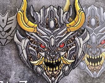 May include: Embroidered patch featuring a detailed robot face with gold horns, red eyes, and a menacing expression. The patch is predominantly gray, with yellow and brown accents. Other robot face patches are visible in the background. The word "Transformers" is at the bottom.