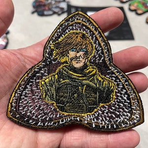 May include: A triangular embroidered patch featuring a detailed image of a person with blonde hair and blue eyes, wearing a dark brown and gold outfit. The patch has a dark brown background with a gold and white border.