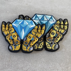 May include: Two yellow robot hands holding a blue diamond-shaped patch. The diamond is surrounded by a yellow border. The robot hands are attached to a blue diamond patch with a yellow border. The patches are sewn together.