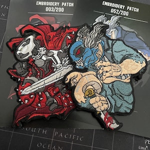 May include: Two embroidered patches with detailed illustrations of action figures. One is in red armor with a sword, the other in blue and tan. The patches are numbered 003/200 and 052/200.