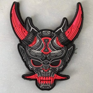 Könnte beinhalten: Ein schwarzer und roter Aufnäher mit einem stilisierten Dämonenmasken-Design. Die Maske hat große Hörner, scharfe Zähne und komplizierte Details in Rot und Grau. Der Aufnäher ist wahrscheinlich aus Gummi oder einem ähnlichen Material.