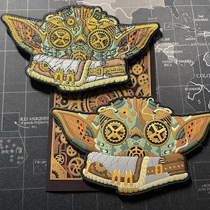 May include: Two embroidered patches featuring a green and brown cartoon character with large ears and a steampunk design. The character has gears and pipes for eyes and a brown leather strap around its neck. The patches are on a brown background with a pattern of gears.