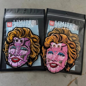 Puede incluir: Dos parches con un retrato estilizado de una mujer con cabello rubio y rasgos faciales robóticos rosas. Los parches están en envases de plástico negro con la inscripción "LIMITED EDITION".