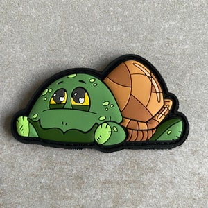 May include: A cartoon turtle patch with a green head and limbs, a brown shell, and a black border. The turtle has large, expressive eyes with heart-shaped pupils.