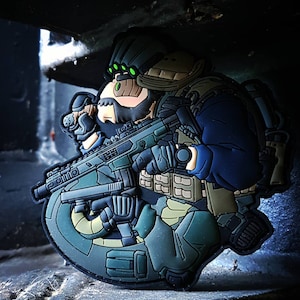 Peut inclure: Un patch détaillé représentant un soldat de style cartoon en équipement tactique, tenant un fusil. Le design incorpore des nuances de bleu, de vert et de beige, avec un dispositif de vision nocturne. Le patch a une base incurvée semi-circulaire.