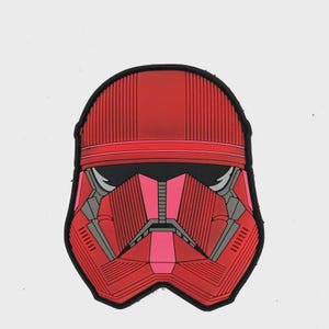 Pode incluir: Um patch vermelho e preto com um design de capacete estilizado. O capacete tem uma placa frontal vermelha e rosa, com detalhes em preto e cinza. O patch tem uma borda preta e é provavelmente um item colecionável ou decorativo.