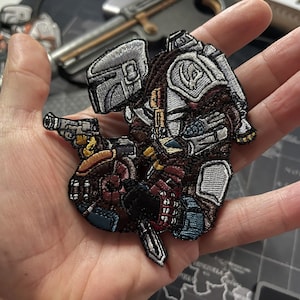 May include: Embroidered patch showing a character in a helmet and armour, holding a blaster. The patch is in shades of brown, grey, and blue, with detailed stitching. The design is likely inspired by a science fiction theme.