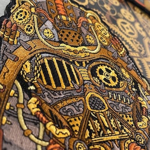 May include: A close-up of a detailed embroidered patch featuring a steampunk-inspired design. The patch is made of fabric and features intricate details of gears, pipes, and other mechanical elements. The patch is primarily gold and black with hints of orange.