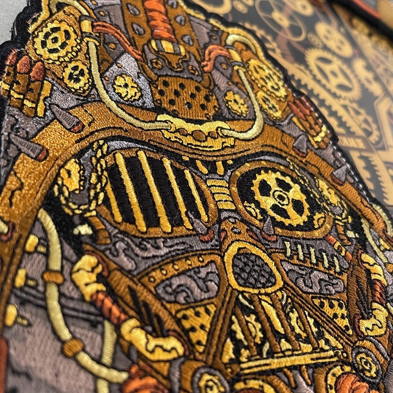 May include: A close-up of a detailed embroidered patch featuring a steampunk-inspired design. The patch is made of fabric and features intricate details of gears, pipes, and other mechanical elements. The patch is primarily gold and black with hints of orange.