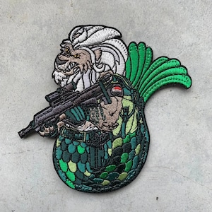 May include: Embroidered patch featuring a mythical creature with a lion's head and a fish tail, holding a rifle. The patch has a green, brown, and white color scheme, with detailed stitching. The design is on a gray background.