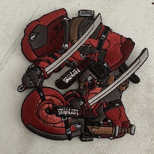 以下が含まれることがあります： 赤いと黒い刺繍のワッペンで、スーパーヒーローのキャラクターが2本の剣を持っている。キャラクターは赤と黒の服を着て、黒いマスクをしている。ワッペンには「GANGL」と書かれている。