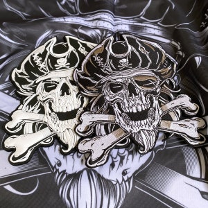 May include: Embroidered patches featuring a skull wearing a pirate hat, crossed bones, and a beard. The patches are black, white, and tan, with detailed stitching. The patches are designed with a pirate theme.