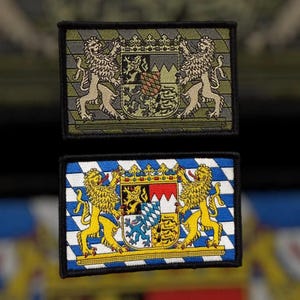 May include: Two embroidered patches featuring the coat of arms of Bavaria. The top patch is on a green camouflage background, while the bottom patch is on a blue and white checkered background.