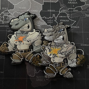 Op de afbeelding: Twee geborduurde patches met cartoon neushoorns die witte shirts en bruine broeken dragen. Beide neushoorns hebben een gele ster op hun borst en zijn vastgebonden met wit touw. De patches zijn geplaatst tegen een grijze kaart achtergrond.