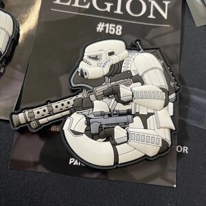 May include: A white and grey patch depicting a Stormtrooper with a blaster rifle. The patch has a black border and the number #158 is visible. The patch is set against a black background.