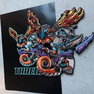 以下が含まれることがあります： 大きな頭、翼、尻尾を持つ漫画の生き物を描いた刺繍パッチが2つ。生き物はヘルメットとゴーグルを着用しています。パッチは、ターコイズ色の「THREAT」という文字が書かれた黒い背景の上にあります。