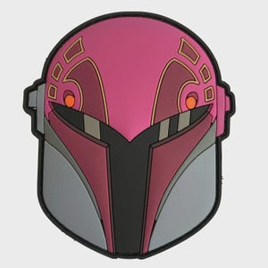 May include: A patch depicting a helmet design in pink, maroon, and grey. The helmet features orange eye details and gold trim. The patch has a black border. The design is a stylized representation.