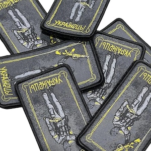 Puede incluir: Un conjunto de parches rectangulares con fondo negro y borde amarillo. Cada parche presenta una ilustración de un soldado y la palabra ucraniana "Українці!" en amarillo. Los parches están apilados.