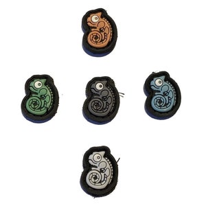 Può includere: Cinque toppe di camaleonte in diversi colori: arancione, verde, grigio, blu e bianco. Ogni toppa è un camaleonte dei cartoni animati con un contorno nero e un occhio bianco.