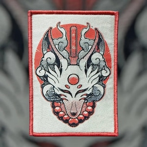 May include: White embroidered patch with a red background featuring a stylised fox face with a red circle in the centre of the face. The fox is surrounded by clouds and a string of beads.