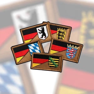Könnte beinhalten: Eine Sammlung von fünf rechteckigen gestickten Aufnähern, die das deutsche Flaggendesign in Kombination mit verschiedenen regionalen Wappen zeigen, darunter ein schwarzer Bär, drei Löwen, ein blau-weißes Rautenmuster, ein roter Löwe und eine grüne Krone.