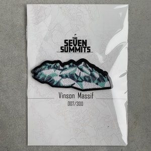 May include: A patch featuring a mountain range design with teal and gray geometric shapes. The patch is labeled "Vinson Massif" and "007/300" below the "The Seven Summits" logo.