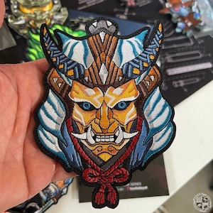 以下が含まれることがあります： 獰猛な表情をした日本の鬼のマスクが描かれたカラフルな刺繍パッチ。マスクは、白と青のパターンで飾られた背景、赤と白のロープ、鋭い牙のペアなど、複雑なディテールで飾られています。