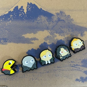 May include: A collection of five cartoon character charms. The charms include a yellow Pac-Man with a wand, a gray Voldemort, a blonde Draco Malfoy, a dark-haired Severus Snape, and a blonde Dobby. The charms are on a brown and blue patterned surface.